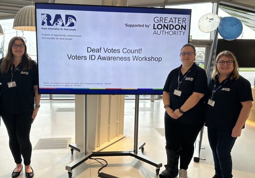 Three RAD staff members standing in front of a large screen that displays the words 'Deaf Votes Count! Voter ID Awareness Workshop'